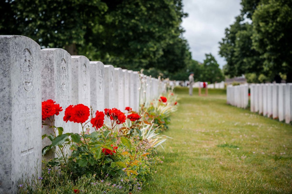 Row of headstones in a CWGC cemetery with bright red roses blooming in the grave borders. In the background, out of focus, can be seen a British soldier standing next to a Union Jack-draped coffn.