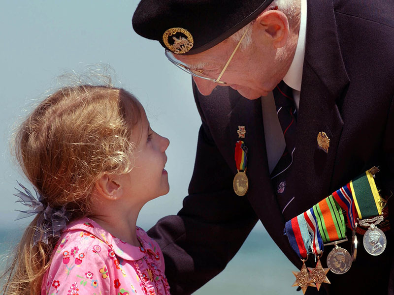 Elderly WW2 veteran in close conversation with a small girl in a pink dress. The veteran is wearing a military beret with cap badge,  a white shirt and a blue blazer with a bar of military medals.