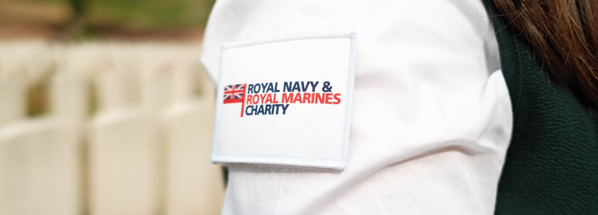 Close up of a white Royal Navy & Royal Marines Charity patch on the arm of a CWGF Guide. The patch shows a Union Jack tied to a red flagpole running parrallel to the text.