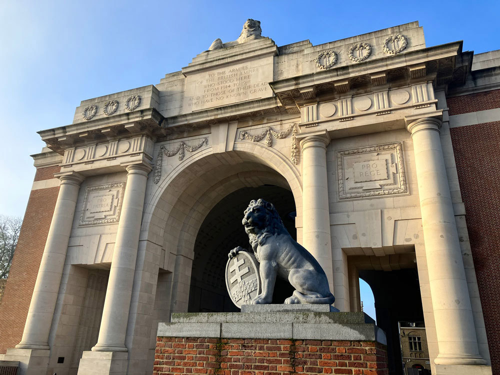 The Menin Gate showing its triumphal arch with one of the lion sculptures in the foreground. The lion is holding a shield in its paws.