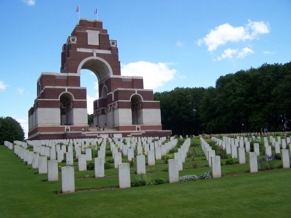 Thiepval Memorial on a summer's day with blue skies contrasting with emerald green grass, white headstones, and ruddy brickwork of the memorial itself.