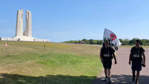 Two young men walk on the Vimy Ridge on a hot summer's day. The Vimy Memorial is visible in the background. The man on the left is holding a white and red poppy flag.