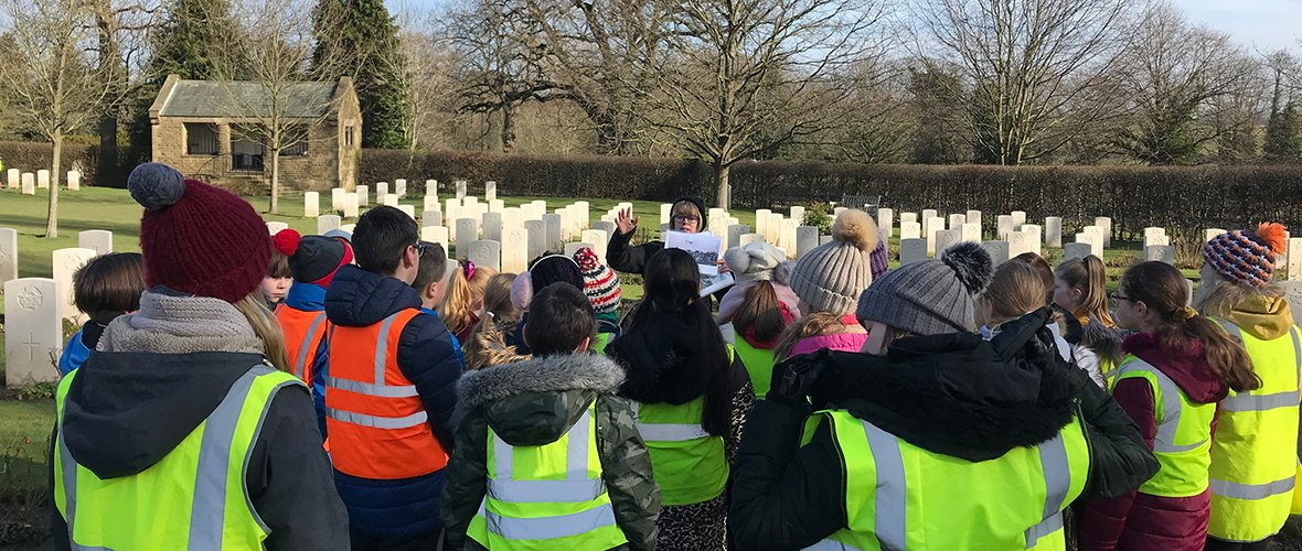 Tour guide gives a talk to schoolchildren in a CWGC cemetery. The children are wearing hi-vis jackets.