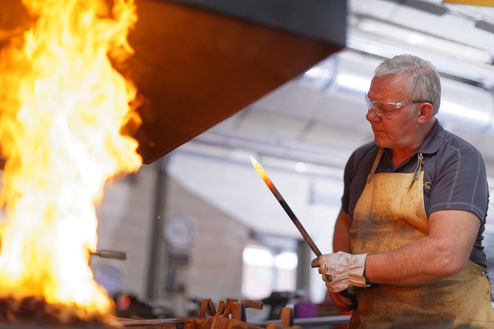 A CWGF blacksmith handles a red hot poker in front of a wild flame in his forge.