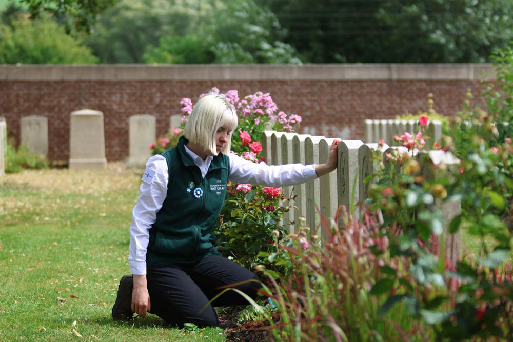 A young women with blonde bobbed hair in a green vest kneels and rests an arm on a CWGC headstone to pay her respects