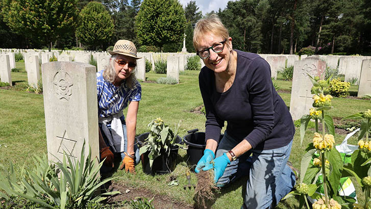 Two kneeling women smile at the camera while trimming the plants and weeds around a CWGC war grave.