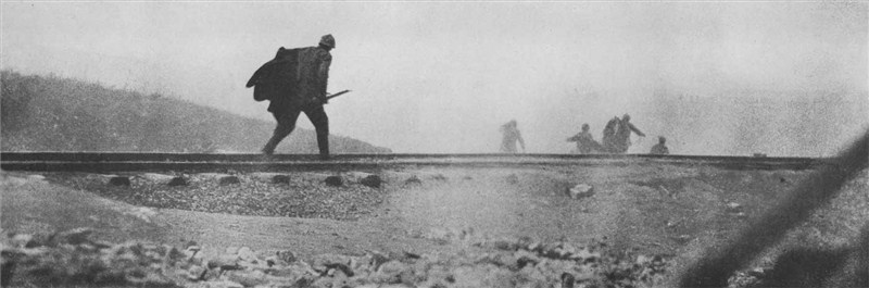 A French soldier in helmet and smock advances across a barren battlefield. His comrades can be seen in the background. The foreground is a morass of churned muddy ground.