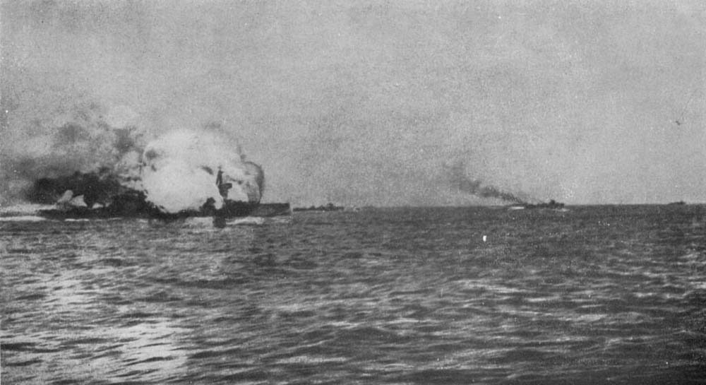 Black and white image of HMS Invincible blowing up during the Battle of Jutland. Smoke billows from the stricken ship which sails on slate grey seas.