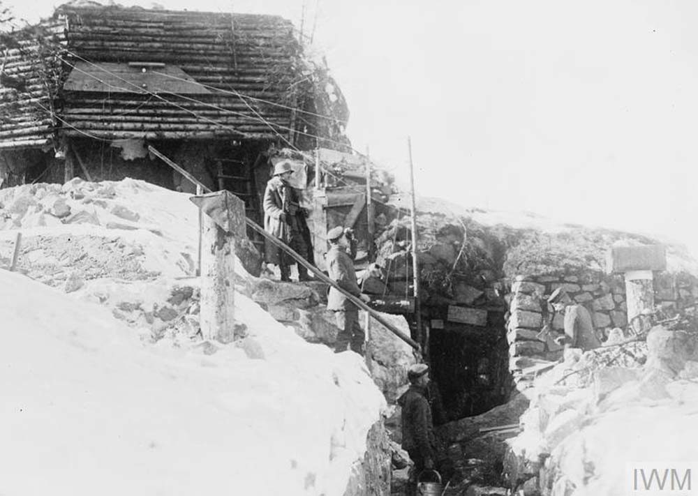 German soldier stands in front a mixed wooden and concrete observation post. The post is accessible via a deep trench which has been reinforced by sandbags.