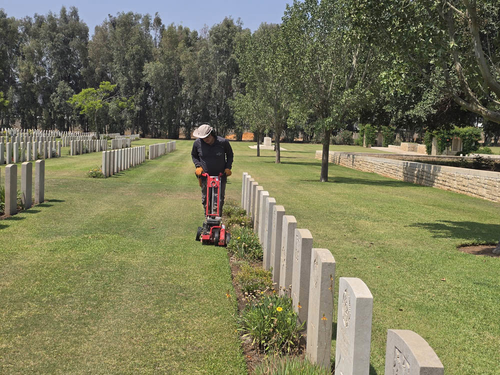 A CWGC Gardener cutting grass with a small, red lanwmower. He is cutting grass next to the boarder of a row of CWGC headstones. Further headstone rows are visible in the background.