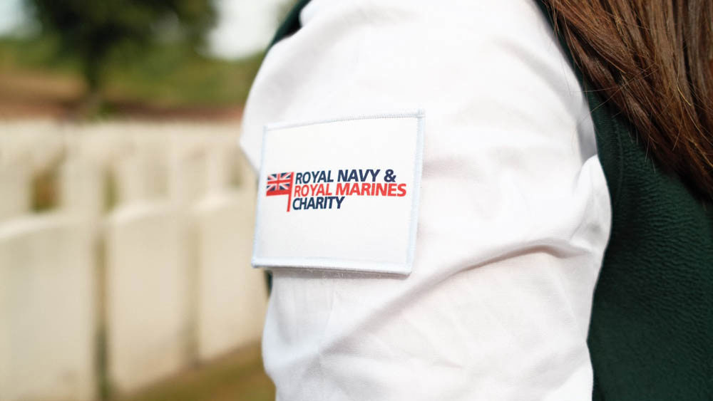 Close up of a white Royal Navy & Royal Marines Charity patch on the arm of a CWGF Guide. The patch shows a Union Jack tied to a red flagpole running parrallel to the text.